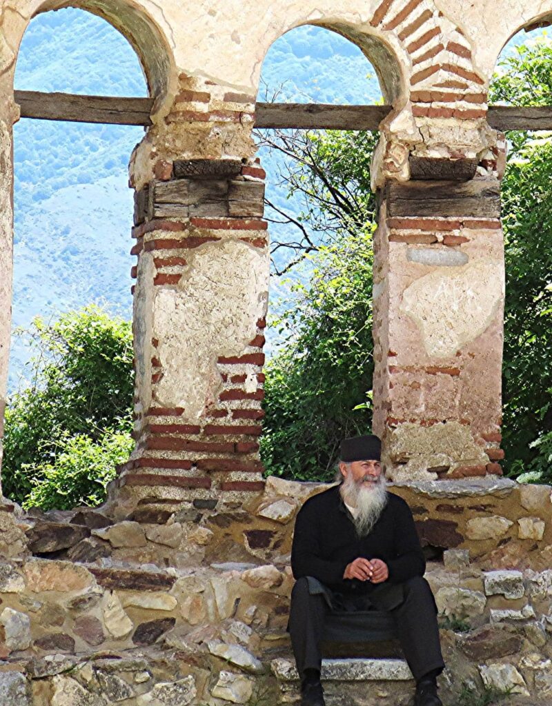 Priest in traditional black clothes with beard seated among ruins of Aghios Achilleos church