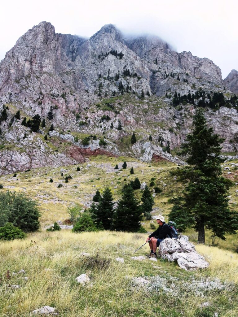 E4 trail below the sheer Mount Vardhousia peaks, with hiker sitting on a rock with grass and scattered pines.